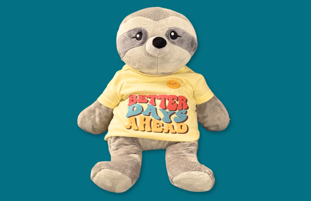 This image, on a dark teal background, shows Sam the Sloth Hugimal wearing a light yellow t-shirt that says Better Days Ahead on it in colorful lettering (orange, teal, dark yellow) and also shows a smiling sun atop the lettering. 