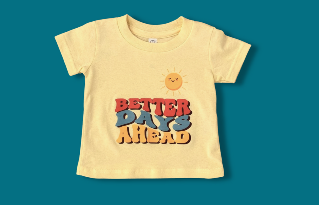 This image, on a dark teal background, shows a light yellow t-shirt that says Better Days Ahead on it in colorful lettering (orange, teal, dark yellow) and also shows a smiling sun atop the lettering. 