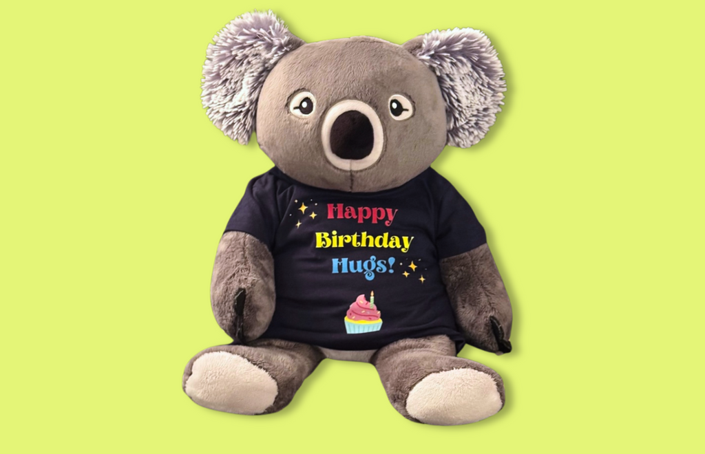 This image shows Quinn the Koala Hugimal wearing a tee-shirt. The shirt is navy blue with red, yellow and blue lettering that says Happy Birthday Hugs, surrounded by yellow sparkle emojis, with a colorful drawing of a cupcake below the lettering. The image is on a highlighter green background