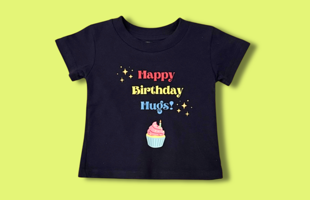 Image shows navy blue t-shirt with red, yellow and blue lettering that says Happy Birthday Hugs, surrounded by yellow sparkle emojis, with a colorful drawing of a cupcake below the lettering. The shirt is shown on a highlighter green background