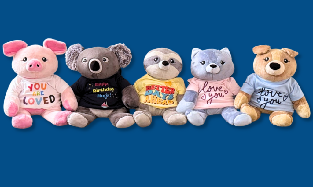 This image shows five Hugimals wearing t-shirts with messages on them, on a dark blue background, arms interlaced. From left: Harper the Pig with a white tee with colorful lettering that says You Are Loved. Quinn the Koala in a navy tee with colorful lettering that says Happy Birthday Hugs. Sam the Sloth in a yellow tee with colorful lettering that says Better Days Ahead. Frankie the Cat in a Pink tee with pink lettering that says Love You. Charlie the Puppy in a light blue tee that says Love You in blue