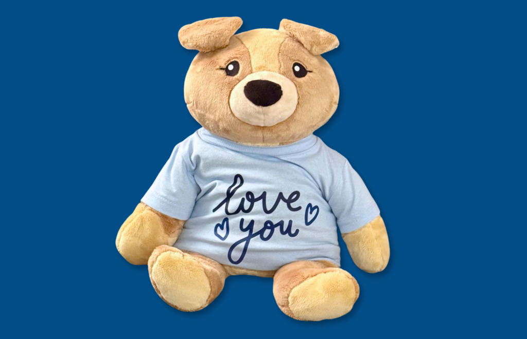 Image of Charlie the Puppy Hugimal on dark blue background. Charlie is wearing a light blue t-shirt with dark blue lettering that says Love You and has two little hearts.