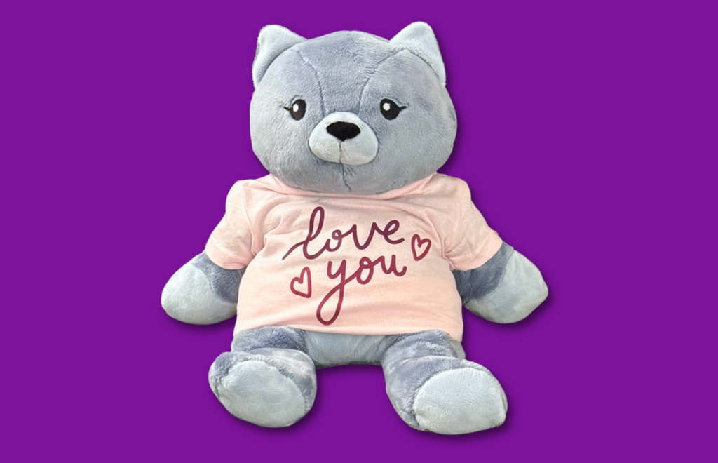 Image shows Frankie the Cat Hugimal on a dark pinkish-purple background wearing a light pink tee with dark pink lettering that says Love You with two little heart outlines.