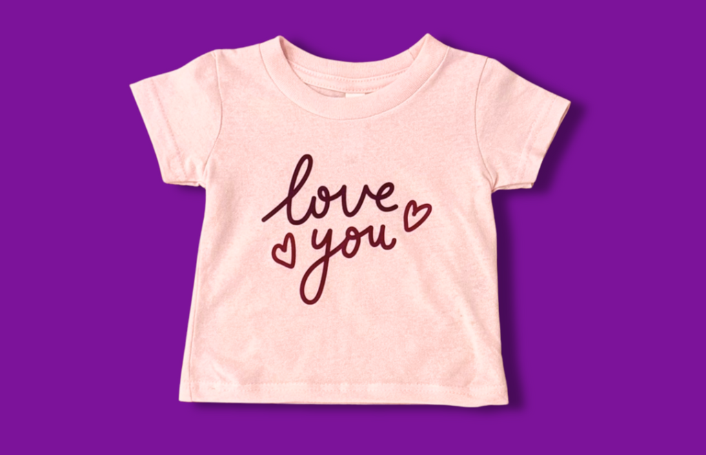 Image shows a light pink tee with dark pink lettering that says Love You and has two little dark pink heart outlines. Background is dark pinkish purple