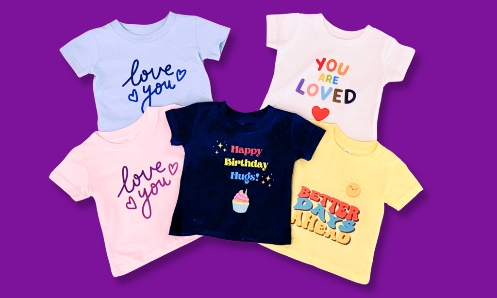 This image, on a dark pinky-purple background, shows five baby tees with messages on them. Clockwise from left: Love You Tee (Blue), You Are Loved (White), Better Days Ahead (yellow), Happy Birthday Hugs (navy), Love You (Pink).