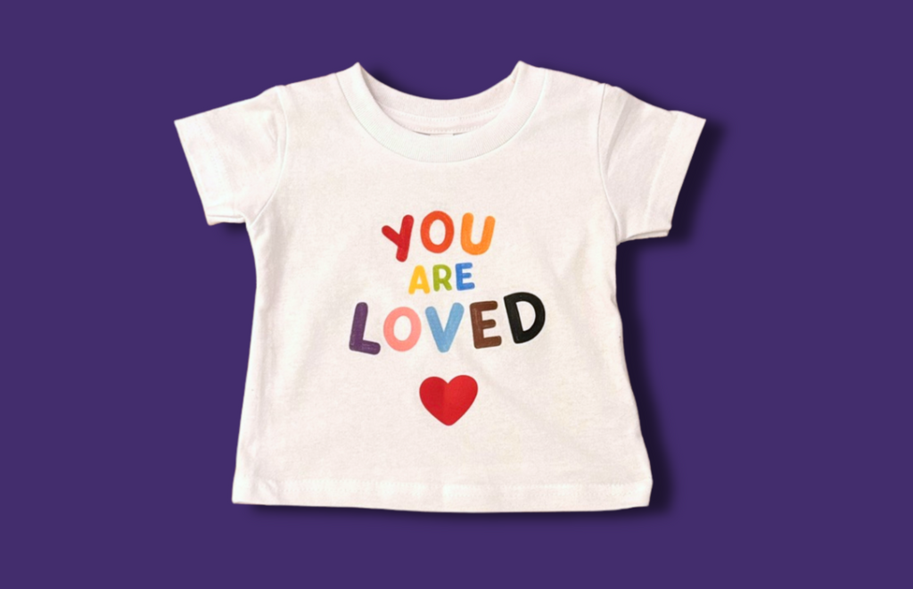 This image shows a white baby tee on a dark purple background. The tee shirt has colorful lettering (red, orange, pink, purple, blue, brown) that says YOU ARE LOVED, with a red heart underneath the lettering.