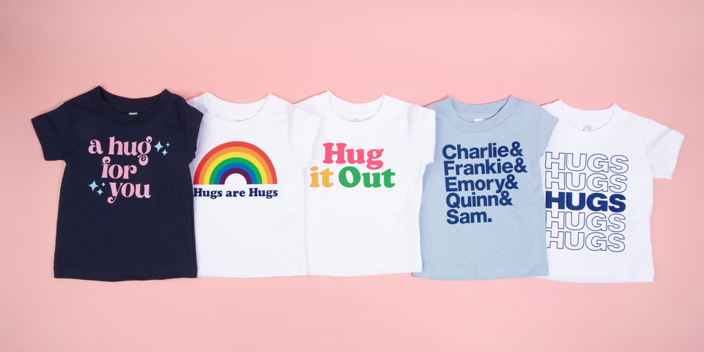 This image shows five baby t-shirts (navy, light blue, and white), spread out on a pink background. The shirts say: A Hug For You, Hugs Are Hugs, Hug It Out, Charlie & Frankie & Emory & Quinn & Sam, and Hugs Hugs Hugs Hugs